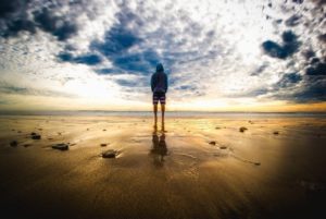 a person standing on the shore looking out at the sea and sunset clouds