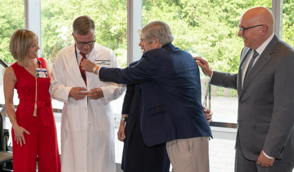 Mr. Schar is helping Dr. O'Connor on with a newly personalized doctor's lab coat.